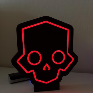 Op de afbeelding: Een zwarte schedelvormige LED-lamp met een rode omtrek. Het licht heeft twee cirkelvormige oogkassen en een driehoekige neus. Het licht staat op een wit oppervlak.
