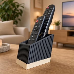 May include: A dark gray remote control holder with a vertical ribbed texture and a solid cream-colored base. The holder contains two black remote controls, one positioned in front of the other, designed to keep electronic controllers organized.