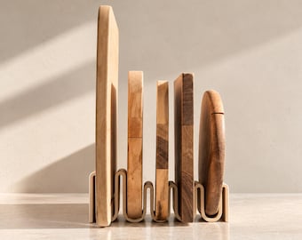 Può includere: Un set di cinque taglieri in legno di diverse altezze e tonalità, tenuti in posizione verticale in un supporto ondulato di colore chiaro. I taglieri presentano venature del legno naturali e bordi lisci e arrotondati.