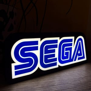 Może przedstawiać: Podświetlany znak SEGA z czarną ramką. Logo SEGA jest niebieskie z białym obrysem. Znak jest podświetlony, tworząc jasny kontrast z ciemnym tłem. Znak umieszczony jest na drewnianej powierzchni.