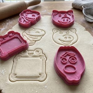 Op de afbeelding: Een set roze plastic koekjesvormpjes gedrukt in een uitgerolde lap deeg. De vormen omvatten een dinosaurus, een varkenskop, een rechthoekig kader met kikkerdetails en een ovaal gezicht met grote ogen en een verbaasde uitdrukking.