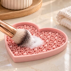 Peut inclure: Un tapis de nettoyage pour pinceaux de maquillage en silicone rose en forme de cœur avec une surface texturée de contours de cœurs en relief. Un pinceau avec une virole en or rose et des poils blancs nettoie sur le tapis avec de la mousse.