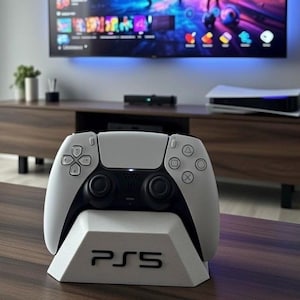Può includere: Un controller PlayStation 5 bianco e nero su un supporto bianco con il logo PS5. Il controller è davanti a una TV e a un mobile di intrattenimento.
