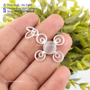 Può includere: Un ciondolo in metallo color argento con una pietra cabochon circolare centrale dall'aspetto pallido e traslucido. La montatura è composta da quattro spirali in filo metallico simmetriche disposte attorno alla pietra, con un occhiello.