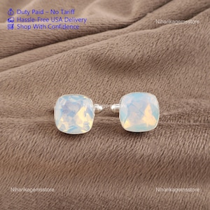 May include: A pair of square cufflinks with iridescent, opalescent stones set in silver. The stones have a faceted appearance, reflecting light. The cufflinks are displayed on a textured, brown surface. The image also includes text about duty and delivery.