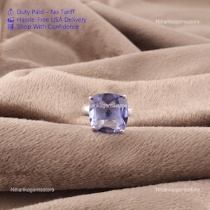 May include: A square-cut, light purple gemstone ring set on a silver band. The ring is displayed on a soft, brown fabric. The text "Duty Paid - No Tariff", "Hassle-Free USA Delivery", and "Shop With Confidence" is visible in the upper left corner.