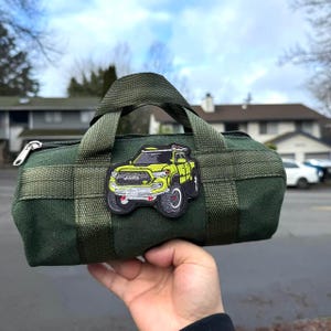 May include: A dark green duffel bag with a woven pattern and a zipper closure. A yellow and black patch of a truck is sewn onto the bag. The bag has a handle and is held up by a person.