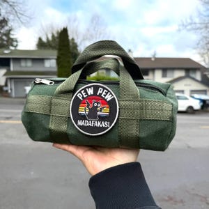 Może przedstawiać: Zielona torba podr&oacute;żna z czarnym zamkiem błyskawicznym i naszywką z napisem "PEW PEW MADAF*KAS!". Na naszywce czarny kot trzymający broń. Torba ma pleciony wz&oacute;r i uchwyt.