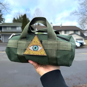 May include: Green canvas bag with a black zipper and a handle. The bag features a patch with a gold triangle and an eye design. The bag is held up in front of a cloudy sky and houses.