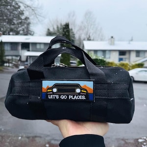 May include: Black duffel bag with a rectangular patch that reads "LET'S GO PLACES." The patch features a car graphic with a mountain and sunset design. The bag has two handles and a zipper closure.