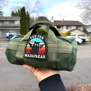 May include: Green canvas duffel bag with a black zipper and handles. The bag features a patch with a black cat holding two guns, the words "PEW PEW" above, and "MADAFAKAS!" below, set against a sunset background.