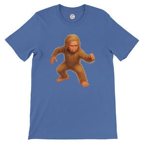 May include: Blue t-shirt featuring a cartoon illustration of a small, furry Bigfoot-like creature. The creature is brown with a stern expression and is in a running pose. The shirt has a crew neck and short sleeves.
