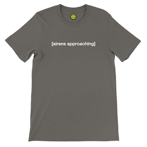 May include: A gray t-shirt with the text "[sirens approaching]" in white. A small yellow smiley face is printed on the upper chest area. The shirt has short sleeves and a crew neck.