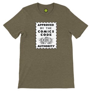 Peut inclure: T-shirt vert olive avec un motif de timbre noir et blanc indiquant "APPROVED BY THE COMICS CODE AUTHORITY". Le t-shirt a un col rond et des manches courtes. Un petit smiley jaune est en haut à droite.