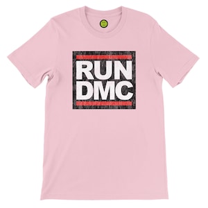 May include: A light pink t-shirt featuring the text "RUN DMC" in a black and white design. The text is inside a black rectangle with red horizontal lines. A small yellow smiley face logo is at the top.