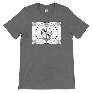 May include: Dark gray t-shirt featuring a black and white test pattern graphic on the front. The graphic includes a circular design with a central emblem and various shapes and lines. The shirt has short sleeves and a crew neck.
