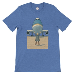May include: Blue t-shirt featuring a graphic of a person lifting a large airplane. The illustration shows the plane's front with the person's arms raised, appearing to hold it up. The shirt has a crew neck and short sleeves.