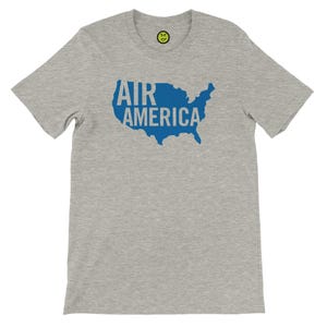 May include: A heather gray t-shirt featuring a blue outline of the United States with the words "AIR AMERICA" printed inside. A small yellow smiley face is visible near the neckline.