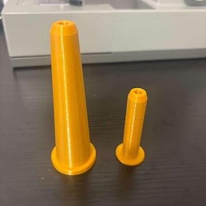 Op de afbeelding: Twee oranje, kegelvormige spoelhouders met een gladde afwerking. De grotere spoelhouder is langer en breder aan de basis, terwijl de kleinere spoelhouder korter en smaller is. Beide voorwerpen hebben een platte, ronde voet en een taps toelopende bovenkant.