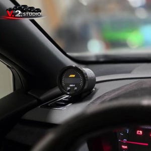 May include: A black digital gauge displaying the text "AEM" and "OBD-II". The gauge is mounted on a black dashboard, with the steering wheel and other car interior elements visible in the background. The gauge is likely for automotive use.