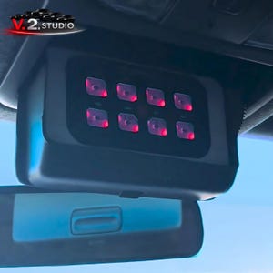 Puede incluir: Un dispositivo electrónico negro con ocho luces cuadradas rojas iluminadas, montado en el techo de un coche. El dispositivo está situado encima de un espejo retrovisor, con el logotipo de V.2 Studio visible en la esquina superior izquierda.