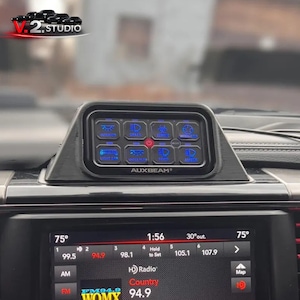 May include: Black dashboard-mounted control panel with illuminated blue icons for various vehicle functions. The panel, branded "AUXBEAM", includes controls for interior lights, grille lights, and more. The vehicle's radio display shows the time, temperature, and radio station.