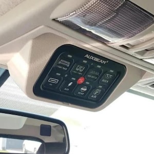 Może przedstawiać: Czarny panel przełączników Auxbeam zamontowany na beżowej konsoli dachowej pojazdu. Panel posiada osiem podświetlanych przycisków z ikonami dla belki świetlnej, kostek świetlnych, świateł dachowych, świateł rock, aux, tylnych i powietrza.