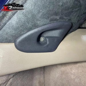 Puede incluir: Primer plano de una palanca de ajuste de asiento de coche de plástico negro con un tornillo plateado. La palanca está sujeta a un material gris oscuro similar al ante y se encuentra sobre una superficie beige. El logotipo V.2 STUDIO es visible en la esquina superior izquierda.