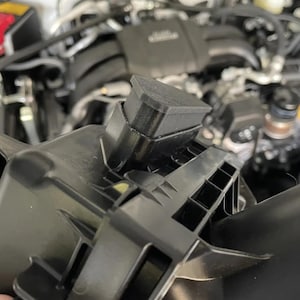 Puede incluir: Un componente automotriz de plástico negro con una tapa superior rectangular y una base moldeada estructurada con soportes laterales acanalados. El objeto se sostiene frente a un compartimento de motor borroso, mostrando su diseño geométrico preciso.