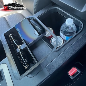 Puede incluir: Un organizador de consola de coche con compartimentos para objetos como una cartera, llaves y gafas de sol. El organizador también contiene dos latas de refresco y una botella de agua. La cartera Ridge es visible.