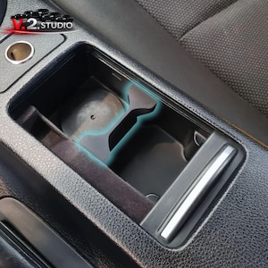 Puede incluir: Compartimento de almacenamiento negro para la consola del coche con un portavasos incorporado y un área de almacenamiento rectangular pequeña. La consola es de plástico negro y tiene una superficie texturizada. El logotipo de V.2 Studio es visible en la esquina superior izquierda.