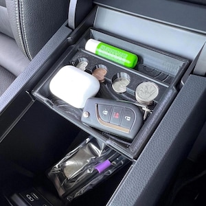 May include: A black 3D-printed car console organizer with compartments for small items. The organizer holds a tube of green lip balm, a pair of white earbuds, coins, and a car key. A small bag with a purple item is in the lower compartment.
