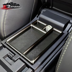 Könnte beinhalten: Schwarzer Auto-Konsolen-Organizer mit einem rechteckigen Fach und zwei kleineren Fächern. Der Organizer hat eine silberne Umrandung und ist für den Einbau in die Mittelkonsole eines Autos konzipiert. Das V.2 Studio-Logo ist sichtbar.