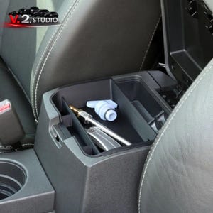 May include: A black car console organizer with a light blue water dispenser spout and a silver metal tool inside. The console is open, revealing storage compartments. The interior is black with white stitching on the surrounding seats.