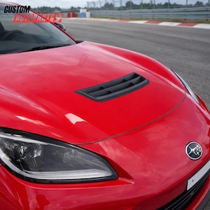 Op de afbeelding: Een felrode motorkap met een zwarte geventileerde luchtinlaat in het midden van het oppervlak. Het voertuig toont een zilveren Subaru-embleem aan de voorzijde en bevat de tekst CUSTOM AUTOPARTS in de linkerbovenhoek van het beeld.