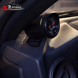 May include: A black gauge with a red needle and white markings is mounted on a car's dashboard. The gauge is set against a black background, with the V.2 Studio logo visible in the upper left corner. The dashboard has a textured surface.