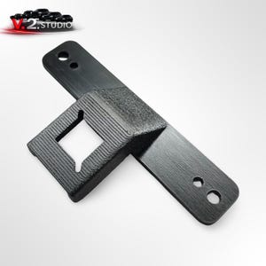 以下が含まれることがあります： 長方形の開口部と2つの円形穴を備えた黒色の3Dプリントブラケット。ブラケットはテクスチャ加工された表面を持ち、オブジェクトを保持するように設計されています。「V.2 STUDIO」のロゴが左上に表示されています。