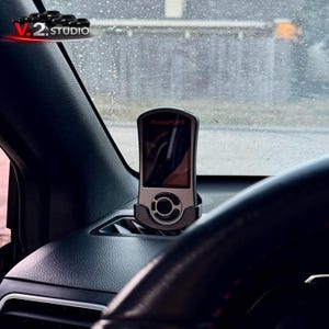 May include: A silver and black Accessport device is mounted on a car's dashboard. The device has a screen displaying information and is held in a black cradle. The car's steering wheel is visible in the foreground, with the V.2 Studio logo in the top left.