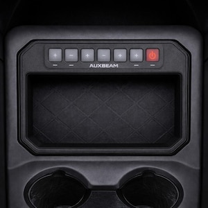 May include: A black automotive center console storage tray featuring a built-in Auxbeam control panel with six buttons marked with plus and minus symbols, a red power button, and a diamond-patterned base, positioned above two circular cup holders.
