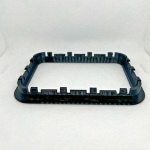 Könnte beinhalten: Ein dunkelgraues, rechteckiges 3D-gedrucktes Objekt mit gezacktem oberem Rand. Das Objekt hat eine quadratische Form mit abgerundeten Ecken und einer soliden Basis. Das Design ähnelt einer Miniatur-Burgmauer oder einem dekorativen Rahmen.