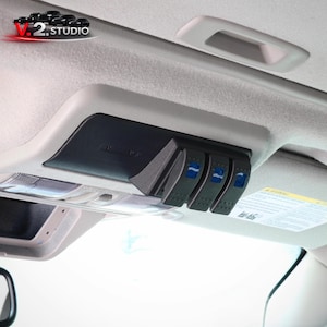 May include: A black car interior console with three blue-accented switches. The console is mounted on a light-colored headliner, with a small storage compartment and a grab handle visible. The V.2 Studio logo is in the upper left corner.