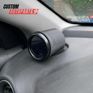Puede incluir: Un soporte de indicador cilíndrico negro montado en un salpicadero de vehículo gris texturizado. El indicador presenta una esfera circular oscura con marcas sutiles y una aguja roja. El texto CUSTOM AUTOPARTS es visible en la esquina superior.