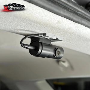 Może przedstawiać: Czarna kamera samochodowa zamontowana na podsufitce pojazdu. Kamera ma cylindryczny korpus, obiektyw i kabel. Uchwyt montażowy jest srebrny. Logo "V.2 STUDIO" jest widoczne w lewym górnym rogu.