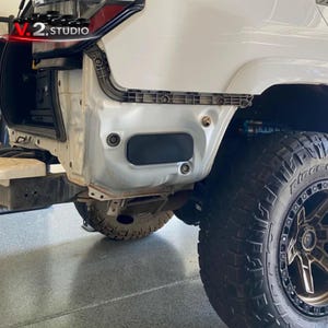 May include: A close-up view of the rear quarter panel of a white vehicle undergoing repair. The panel is partially disassembled, revealing internal components. A black rectangular access panel is visible, along with a large, black off-road tire.