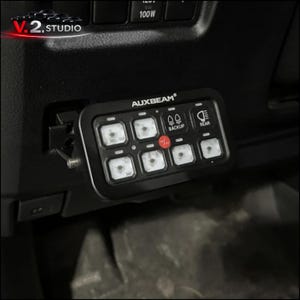 May include: Black AUXBEAM control panel with eight illuminated square buttons, a red button, and icons for backup and rear lights. The panel is mounted on a black surface, likely inside a vehicle.