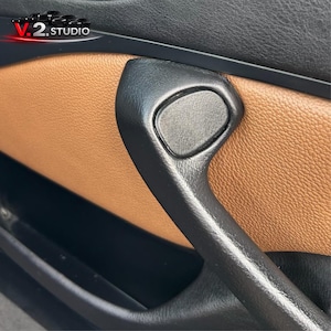 Könnte beinhalten: Nahaufnahme eines Autotürgriffs. Der Griff ist schwarz und nach rechts gebogen, mit einer strukturierten braunen Lederverkleidung im Hintergrund. Die Oberseite der Tür ist schwarz.