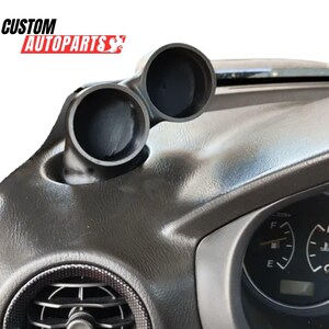 Puede incluir: Un soporte doble para indicadores de color negro instalado en el salpicadero de un vehículo, con dos aberturas circulares para instrumentación. El texto CUSTOM AUTOPARTS es visible en la esquina superior izquierda, con un elemento gráfico.