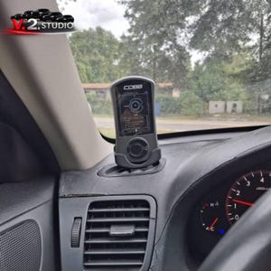 May include: A black COBB tuning device is mounted on a car's dashboard. The device displays information on its screen. The dashboard is black, and the car's interior is visible, including the steering wheel and air vent.