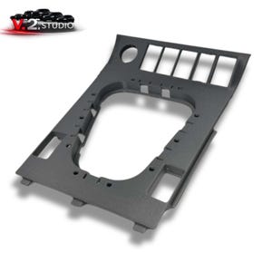 Könnte beinhalten: Ein dunkelgraues Autoinnenkonsolenbauteil mit mehreren rechteckigen Öffnungen und einem kreisförmigen Loch. Das Bauteil hat eine strukturierte Oberfläche und ist für den Einbau in das Armaturenbrett eines Fahrzeugs konzipiert. Das Logo "V.2 Studio" ist sichtbar.