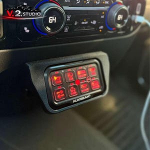 Może przedstawiać: Czarny panel z ośmioma podświetlanymi na czerwono przyciskami, oznaczonymi ikonami i tekstem, w tym "HORN" i "CARGO". Panel jest zamontowany w desce rozdzielczej pojazdu, w pobliżu elementów sterowania klimatyzacją. Widoczna jest nazwa marki "AUXBEAM".